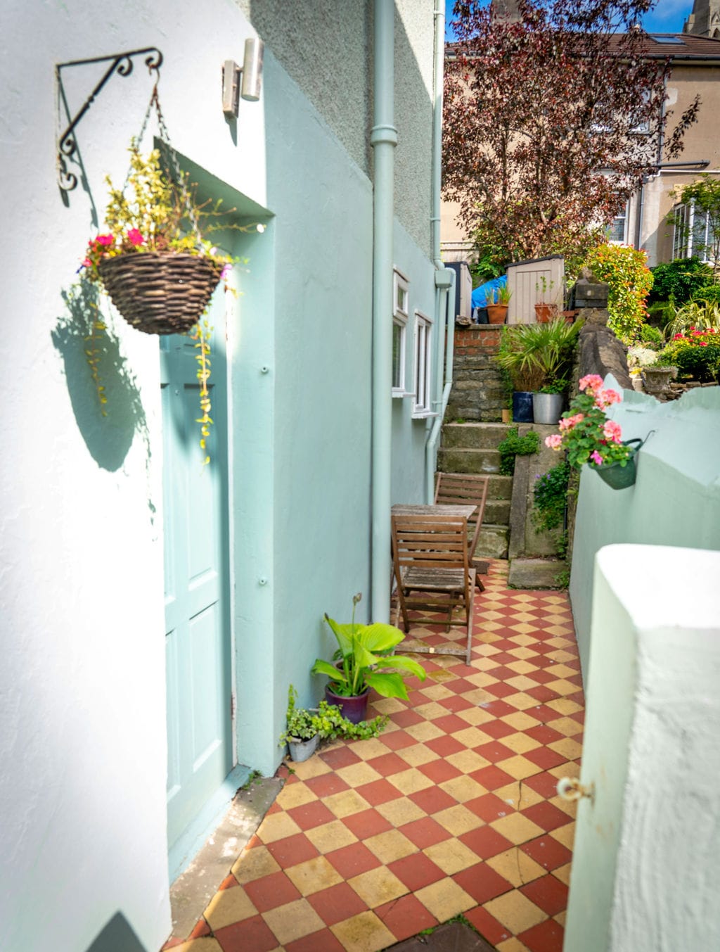 A charming entrance area is visible, featuring a pale blue door framed by greenery and a hanging basket. The pathway showcases a decorative tiled pattern in warm hues, leading past a small seating area with a wooden bench, surrounded by plants and garden elements.
