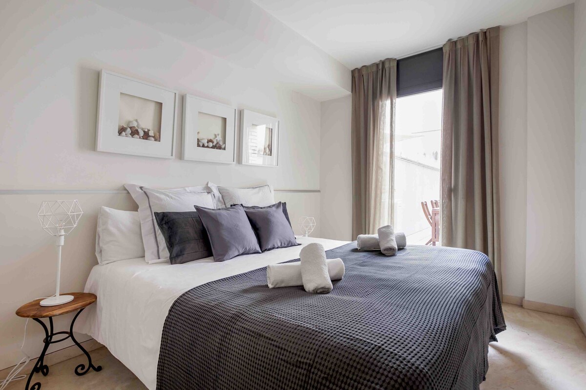 A comfortable bedroom features a spacious bed adorned with gray and white linens. Complementing the decor, three framed artworks are displayed above the bed. Natural light enters through a window dressed with beige curtains, which overlooks a small outdoor seating area.