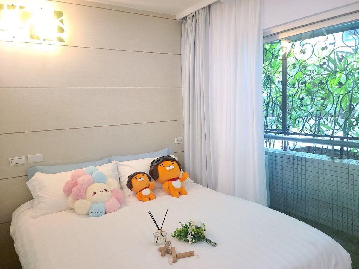 A cozy sleeping area features a spacious bed with light bedding. Two plush toys sit atop colorful cushions. Natural light filters in through sheer curtains, illuminating the room and offering views of greenery outside, complemented by decorative items arranged on the bedside.