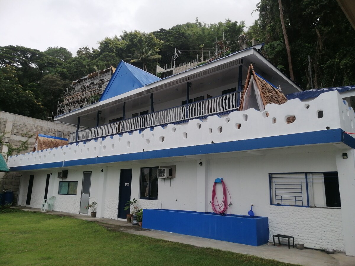 A two-story building features a distinct blue roof and white walls, accented by decorative eaves. The lower level includes windows and air conditioning units, while the upper level is adorned with Kubo huts. Green lawn space surrounds the structure, creating a serene environment.