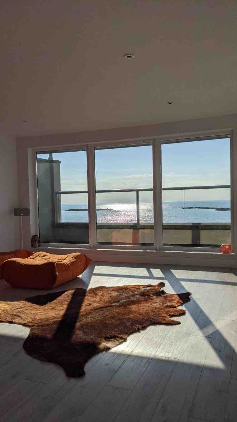 A bright and airy space features large glass windows offering expansive views of the shimmering sea. A cozy orange seating area and a warm, textured rug add comfort, while natural light fills the room, creating a serene atmosphere.
