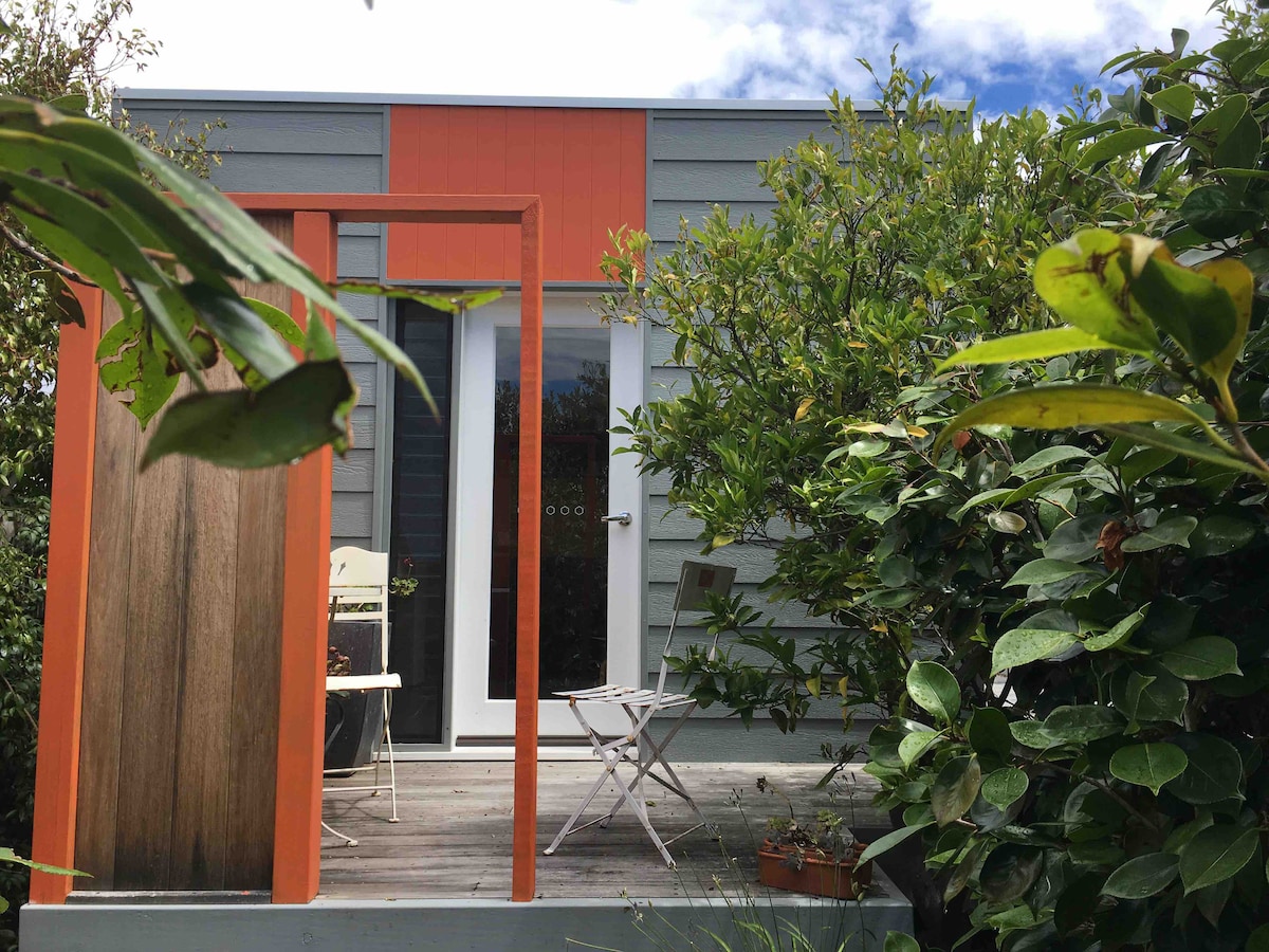 A private entrance to the studio is framed by vibrant greenery, showcasing a small balcony area furnished with two chairs. The entrance features a large glass door, allowing natural light to illuminate the interior space. Freshly renovated exterior details are visible.