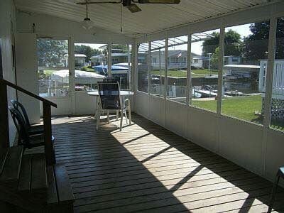 A screened-in porch features wooden flooring and a ceiling fan. A dining table with chairs is positioned near large windows, allowing plenty of natural light. The view outside reveals grassy areas and boats, creating a relaxed environment.