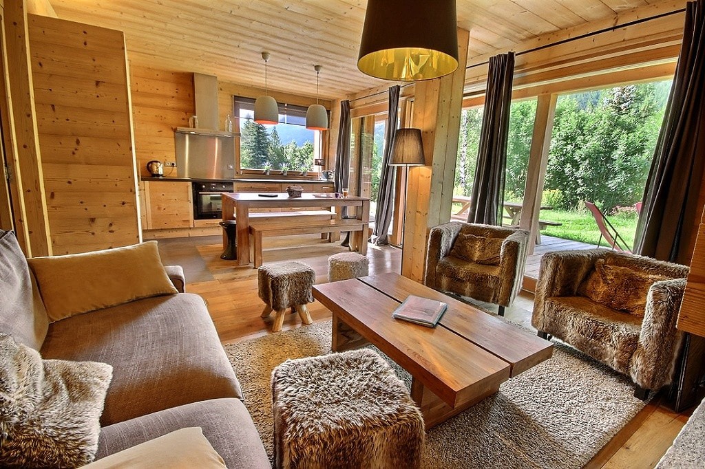 A welcoming living area is designed with natural wood elements. A comfortable sofa and two armchairs are complemented by a wooden coffee table. Large windows allow natural light to illuminate the space, leading to a view of the outdoors. A plush area rug adds warmth.