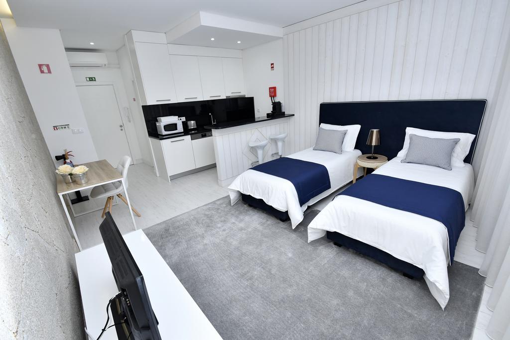 A modern accommodation space features two twin beds with navy blue accents and white linens. A kitchenette is visible in the background, equipped with essential appliances. A small dining table sits nearby, and a neutral-colored rug covers the floor, enhancing the contemporary design.
