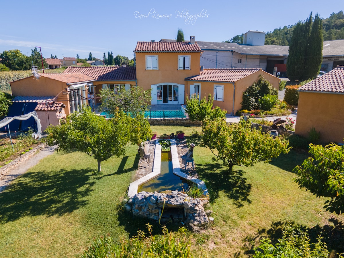 A spacious family home is surrounded by a well-maintained garden. A swimming pool is visible at the rear, complemented by a stone pathway that leads through the lawn. Several trees line the area, providing greenery and shade, while the structure features a warm color palette.