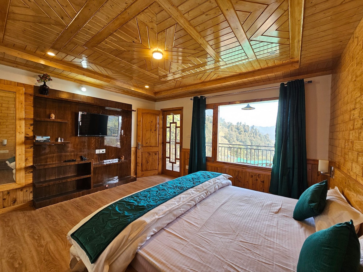 A spacious bedroom features wooden paneling and a high ceiling, complemented by large windows offering scenic views. The room is equipped with a comfortable bed covered in white linens and a turquoise throw. Dark curtains frame the windows, enhancing the natural light that enters.