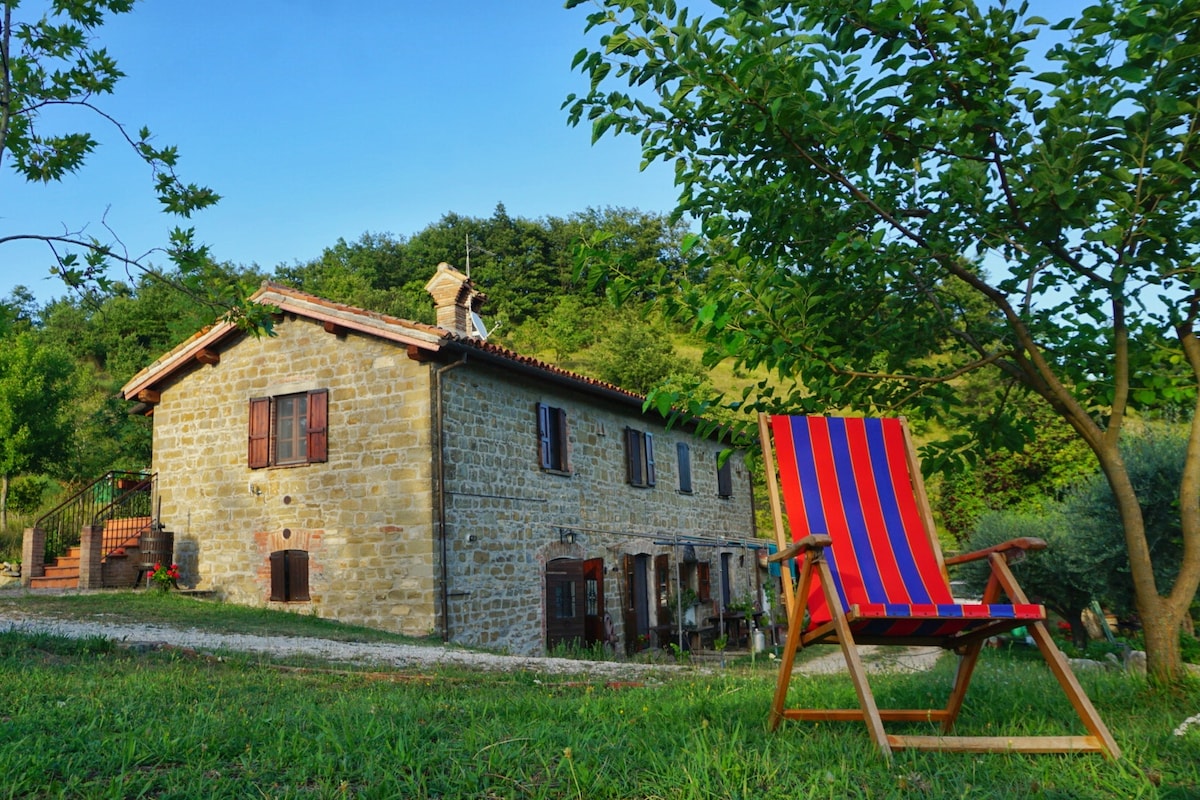 A stone house is set against a backdrop of green hills. A red and blue striped deck chair is positioned on the grass, providing a relaxing spot. The house features wooden windows and a chimney, surrounded by trees and a pathway leading to the entrance.