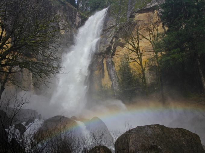 A majestic waterfall cascades down rocky cliffs, creating a mist that generates a rainbow across the bottom. Surrounding greenery and scattered boulders enhance the natural landscape, exemplifying the serene beauty of the area.