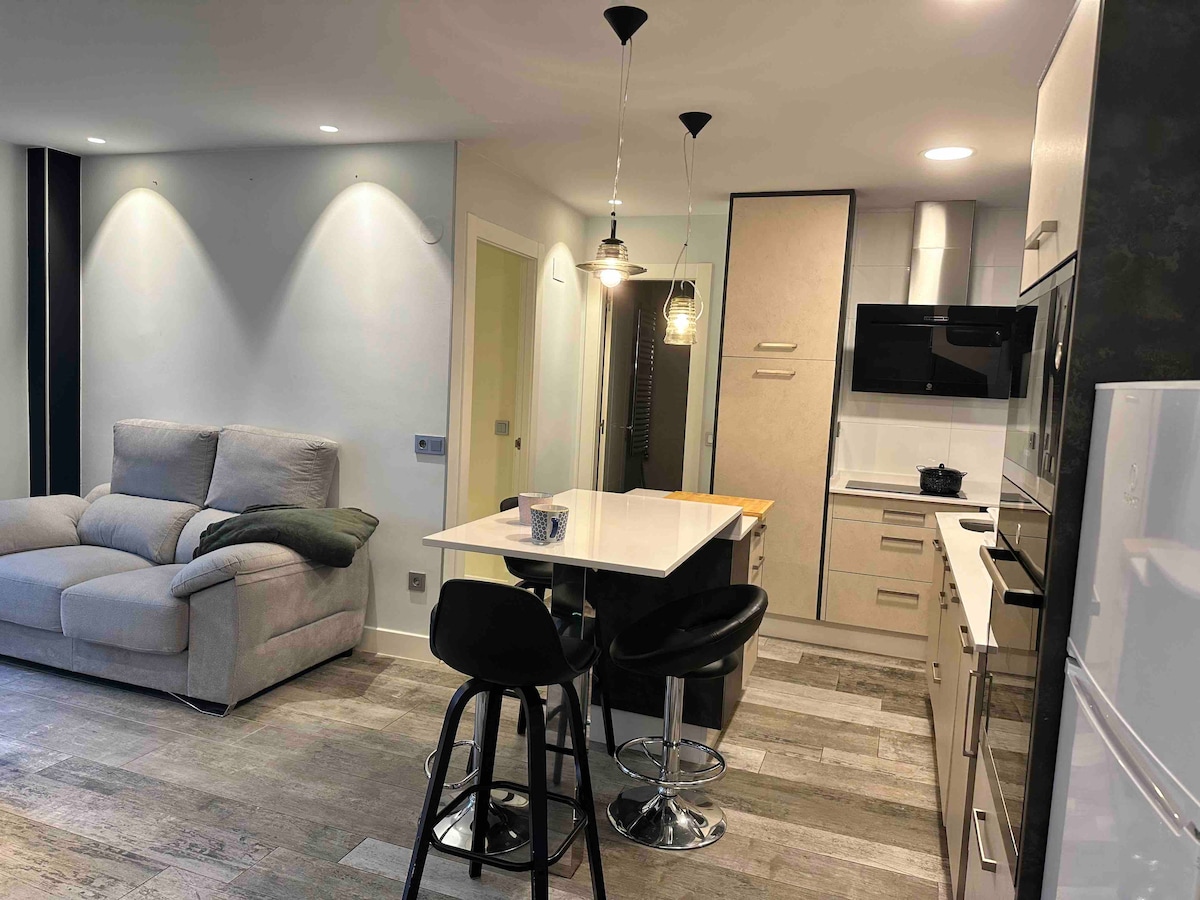 A modern open-plan living area features a neutral-toned sofa and a sleek kitchen with updated appliances. A breakfast bar with black stools is highlighted, while ample lighting illuminates the space, enhancing the contemporary design. Clean lines and a functional layout are evident throughout.