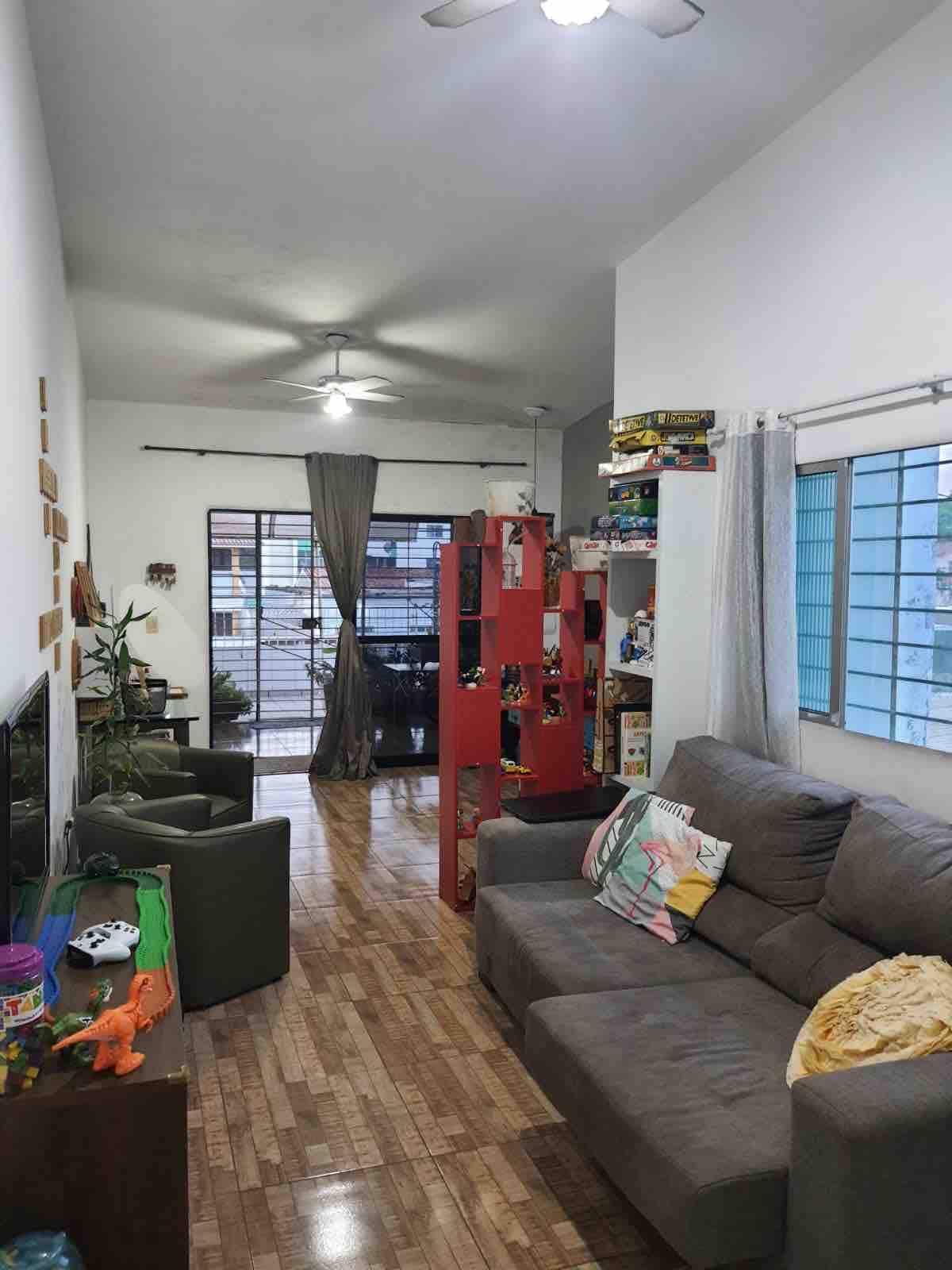 A spacious living area features a gray sofa and two green armchairs, complemented by a vibrant bookshelf filled with games and decor. Natural light filters in through multiple windows, enhancing the room's welcoming ambiance. A small indoor plant is visible near the entrance.