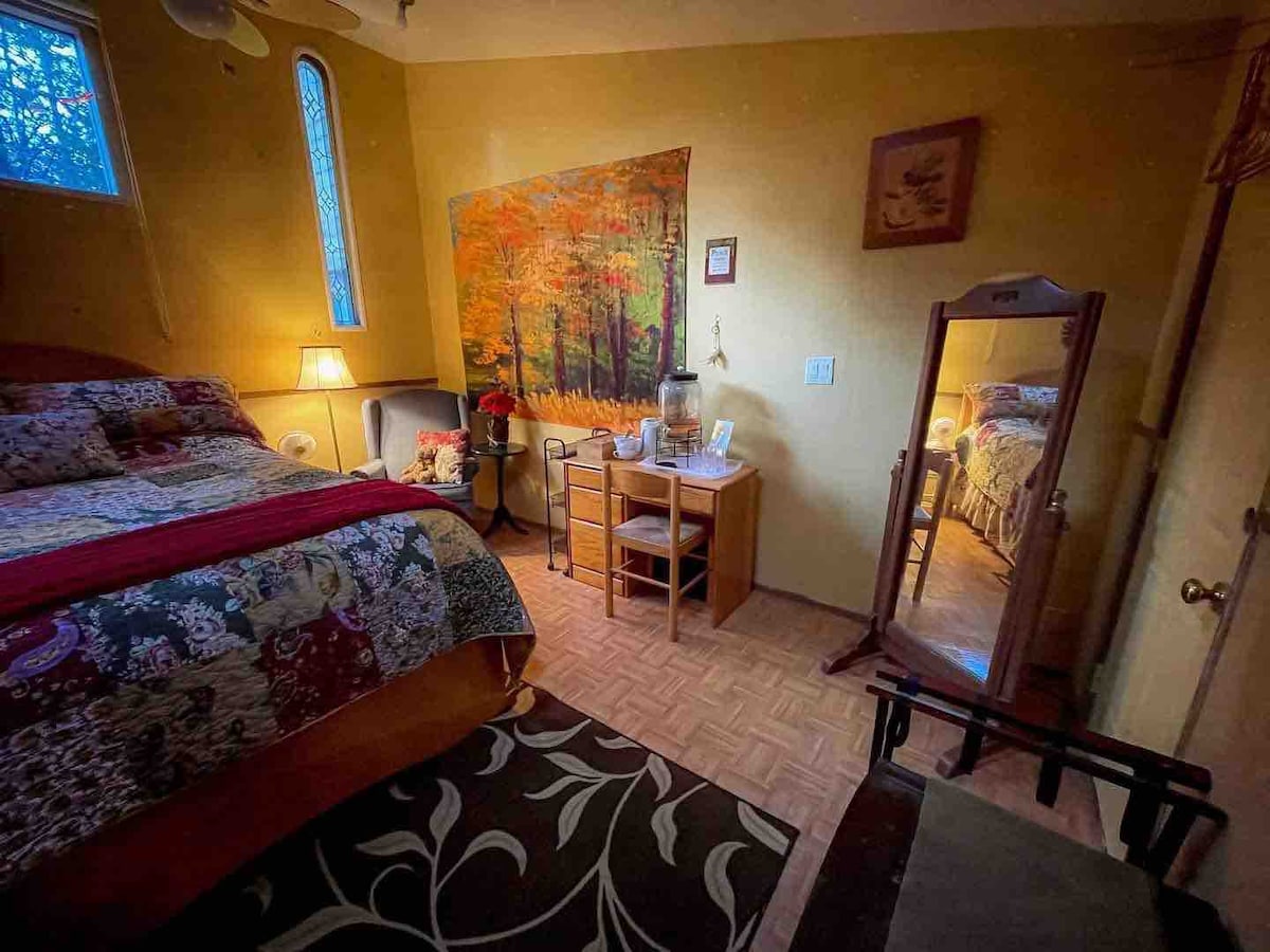 The room is warmly lit, featuring a spacious bed dressed with floral bedding. A nature-themed mural decorates one wall alongside a mirror and wooden furniture, including a small table and chair. A cozy area rug adds texture to the wood flooring.