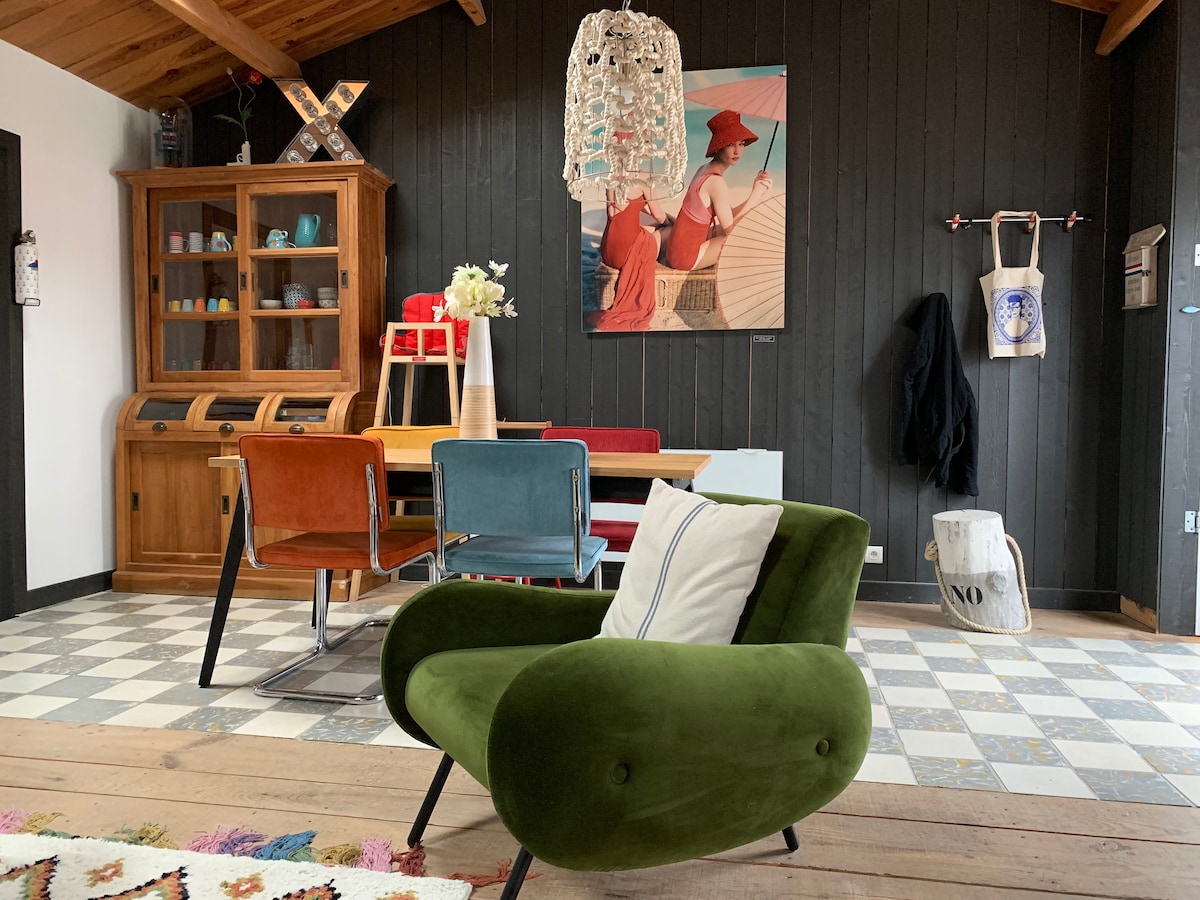 A stylish interior showcases a vibrant green armchair and a wooden dining table surrounded by colorful chairs. A decorative cabinet holds various items, and a modern chandelier hangs from the sloped ceiling. Artwork on the wall features two figures, adding a lively touch to the space.