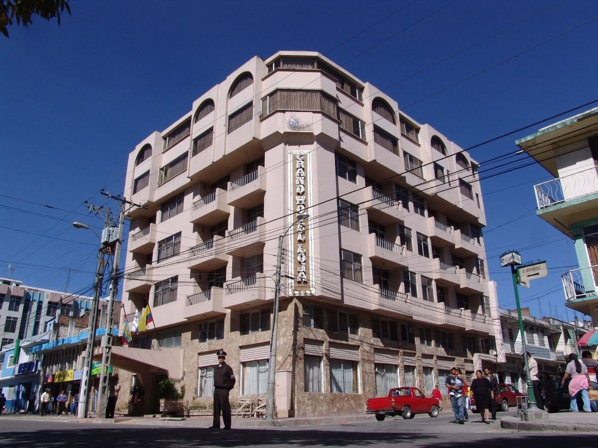 A four-story building is presented with a light-colored façade and large windows. The front features a prominent sign displaying the hotel's name, while street views include parked vehicles and pedestrians, suggesting a vibrant city atmosphere. Nearby shops and utilities are visible in the surroundings.