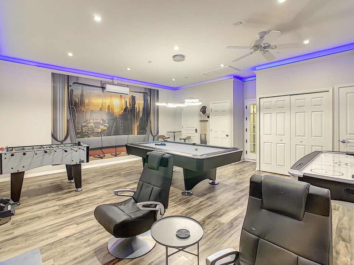 A game room with modern amenities features a full-size pool table, foosball table, and air hockey table. LED lights create an inviting atmosphere. Two gaming chairs are situated near the pool table, and a wall mural of a cityscape adds a dynamic backdrop.