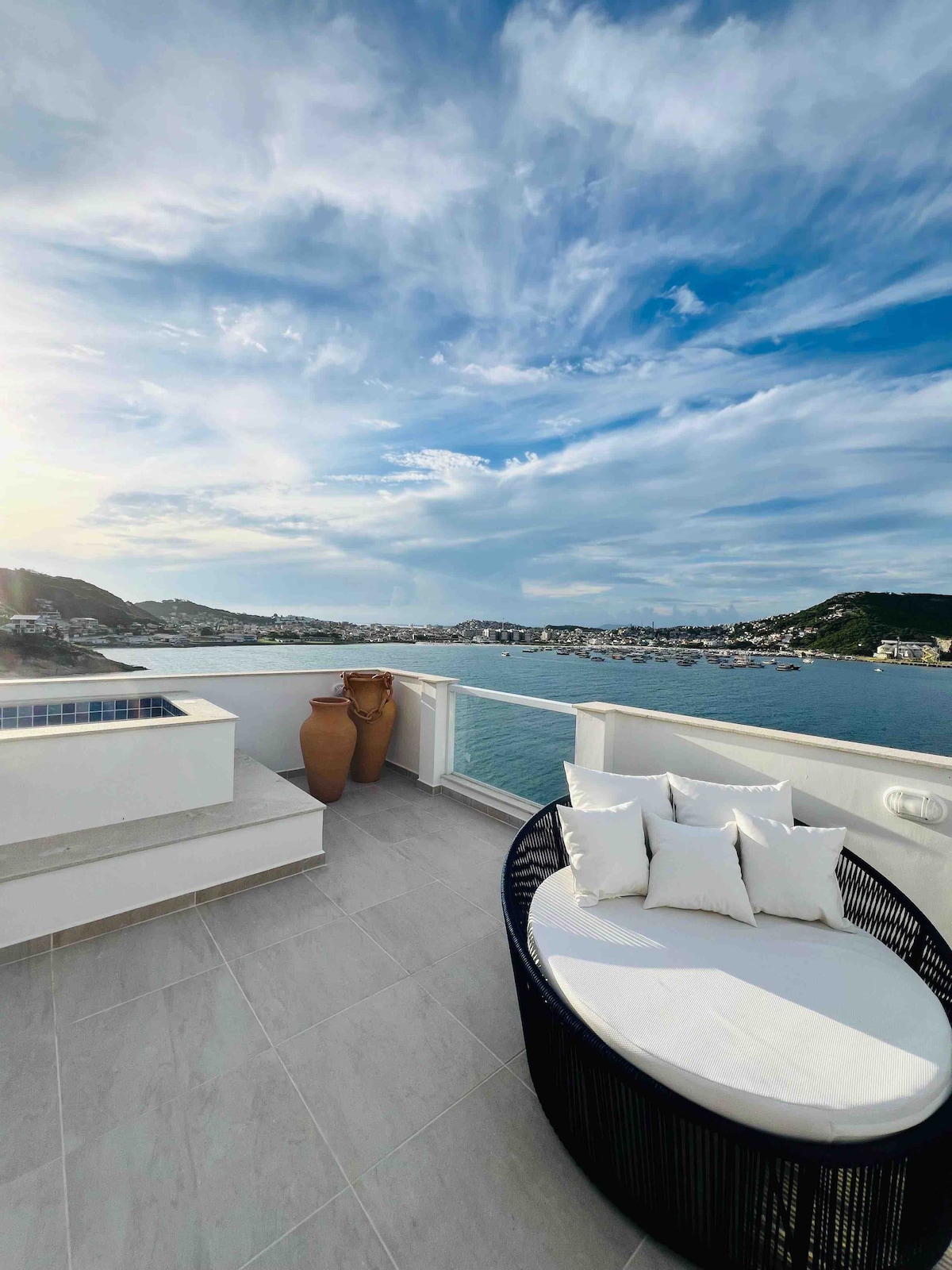 A scenic terrace is showcased, featuring a stylish round seating area adorned with four plush white pillows. Two decorative pots are positioned nearby, while the expansive view of the water and distant hills is framed by a partly cloudy sky.