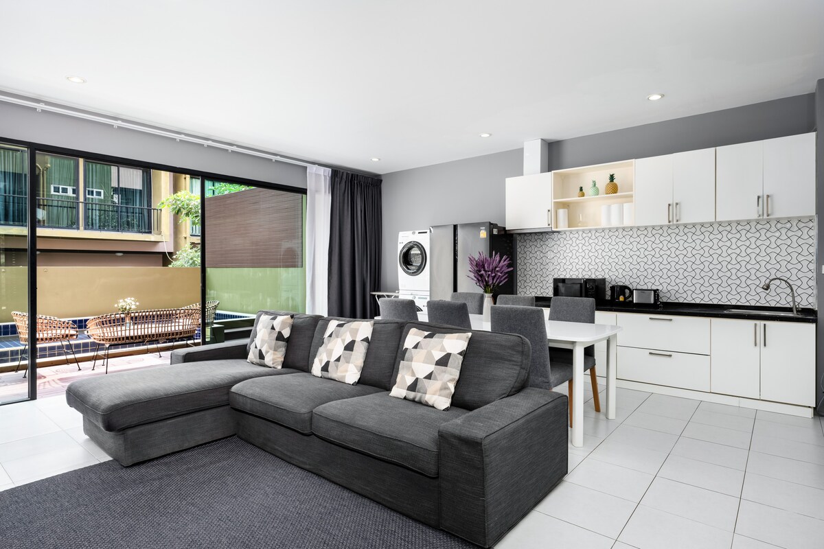A modern living area is displayed with a spacious sectional sofa and patterned cushions. The fully equipped kitchen features sleek cabinetry and a dining space. Large glass doors allow for natural light and access to an outdoor patio with seating.
