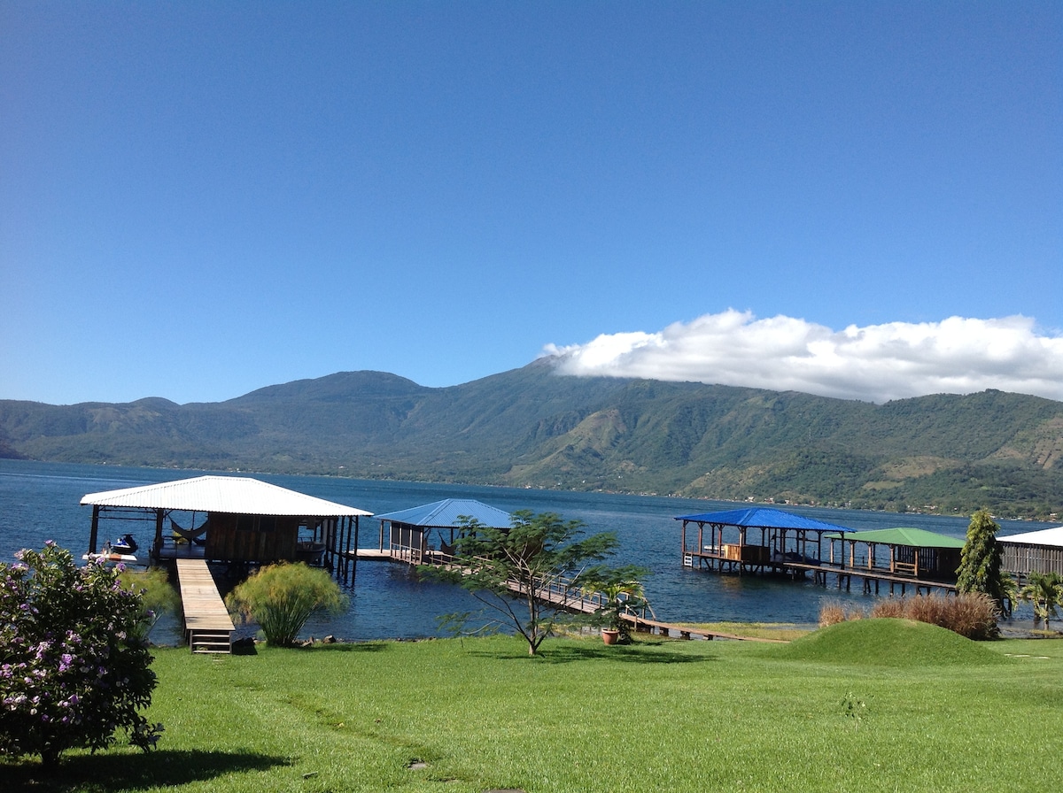 A serene waterfront view showcases large wooden docks extending into calm waters. Lush greenery borders the landscape, with distant mountains providing a picturesque backdrop under a clear blue sky. Light clouds rest softly above the mountains, creating a sense of tranquility.