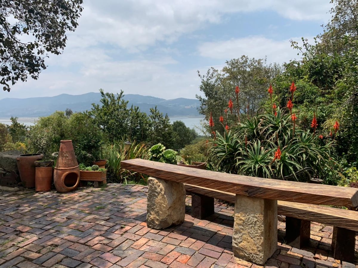 A wooden bench rests on a stone-paved patio, surrounded by vibrant greenery and flowering plants. In the background, a scenic view reveals mountains and a body of water under a cloudy sky.