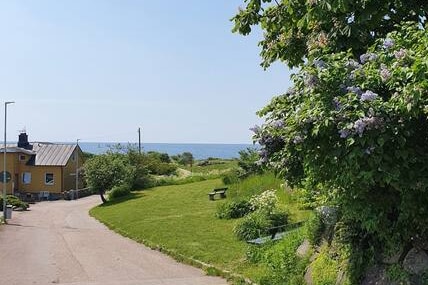 A view of a serene pathway leading towards the sea, surrounded by lush greenery and blooming plants. An inviting yellow building is visible in the background, set among the peaceful landscape, with glimpses of the ocean reflecting the clear blue sky.