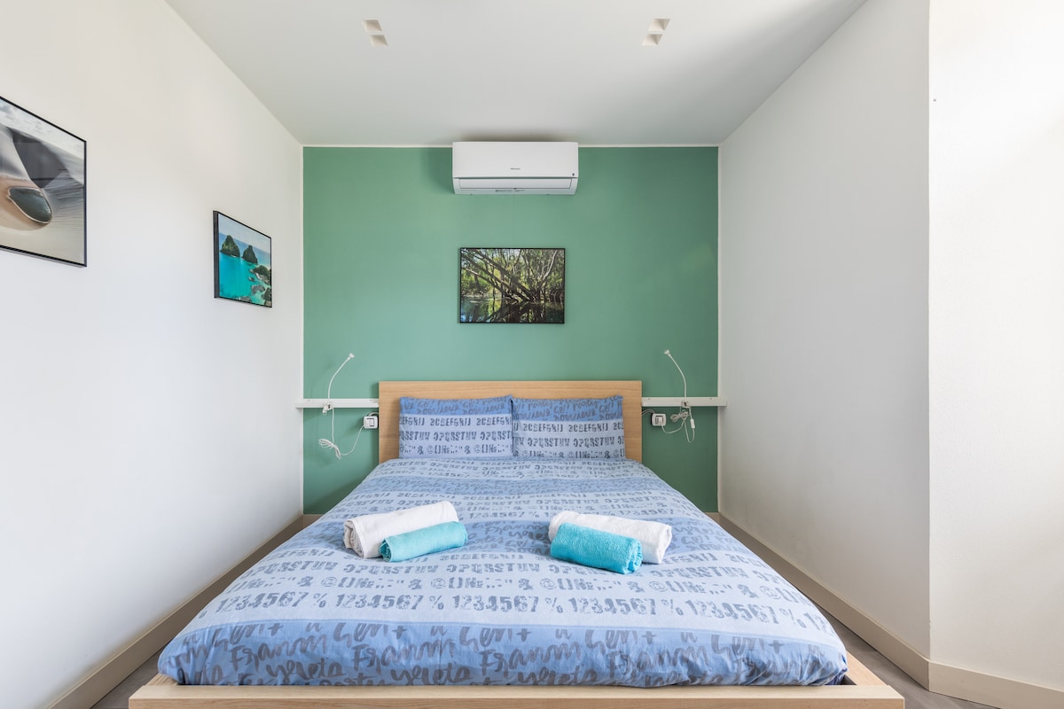 The spacious bedroom features a large bed with a blue patterned cover and towels neatly arranged at the foot. Green accent walls add a calming touch, complemented by artwork showcasing landscapes. An air conditioning unit is mounted above the bed for climate control.