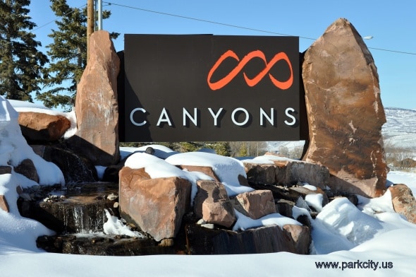 A prominent sign displaying the word 'Canyons' is surrounded by large, natural stone features. Snow blankets the ground, and a small water feature flows nearby, set against a clear blue sky.