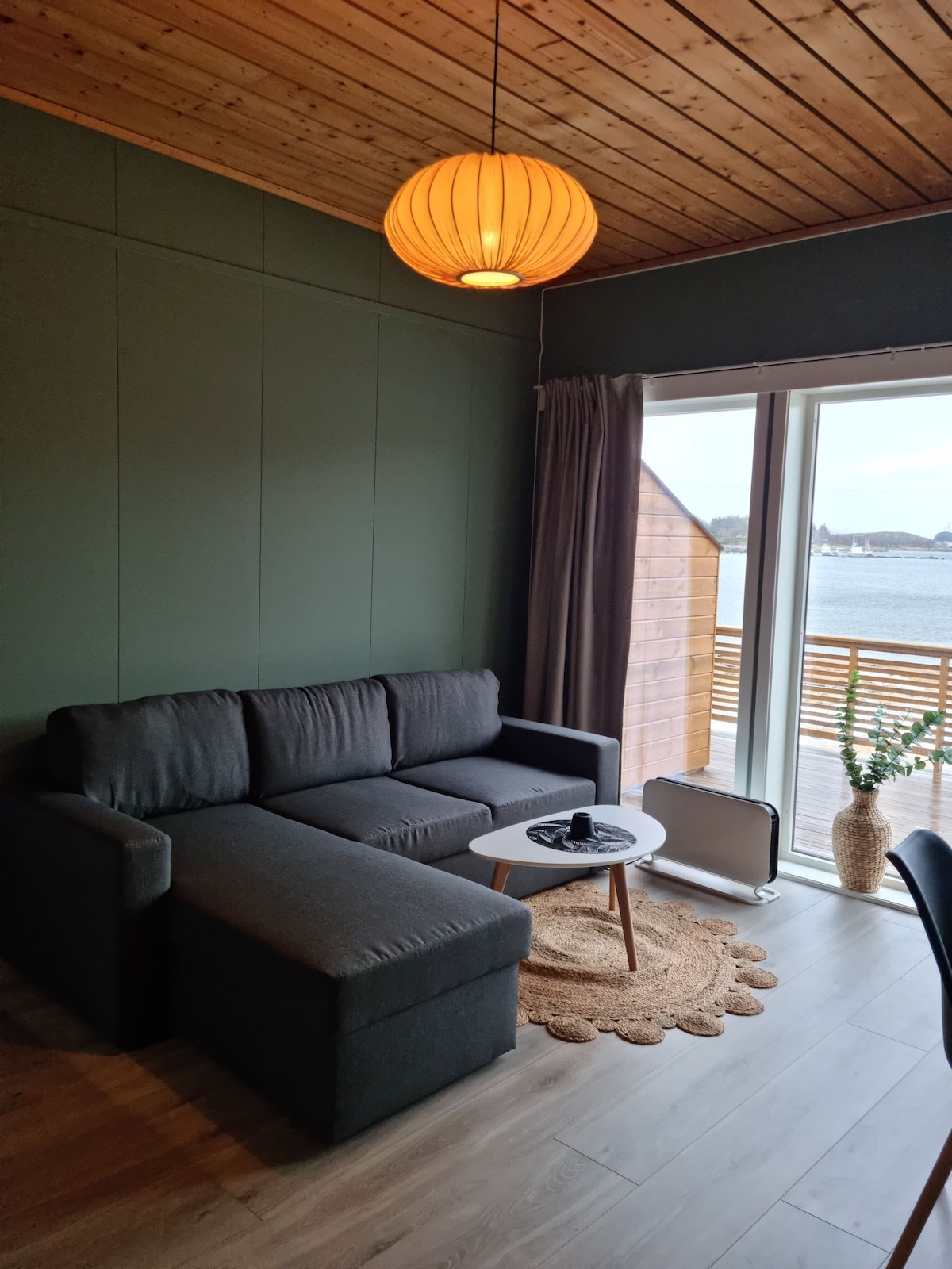 A cozy living area features a dark grey sectional sofa adjacent to a round wooden coffee table. Natural light enters through large sliding doors that open to a scenic waterfront view. A soft area rug complements the space, creating a relaxing atmosphere.