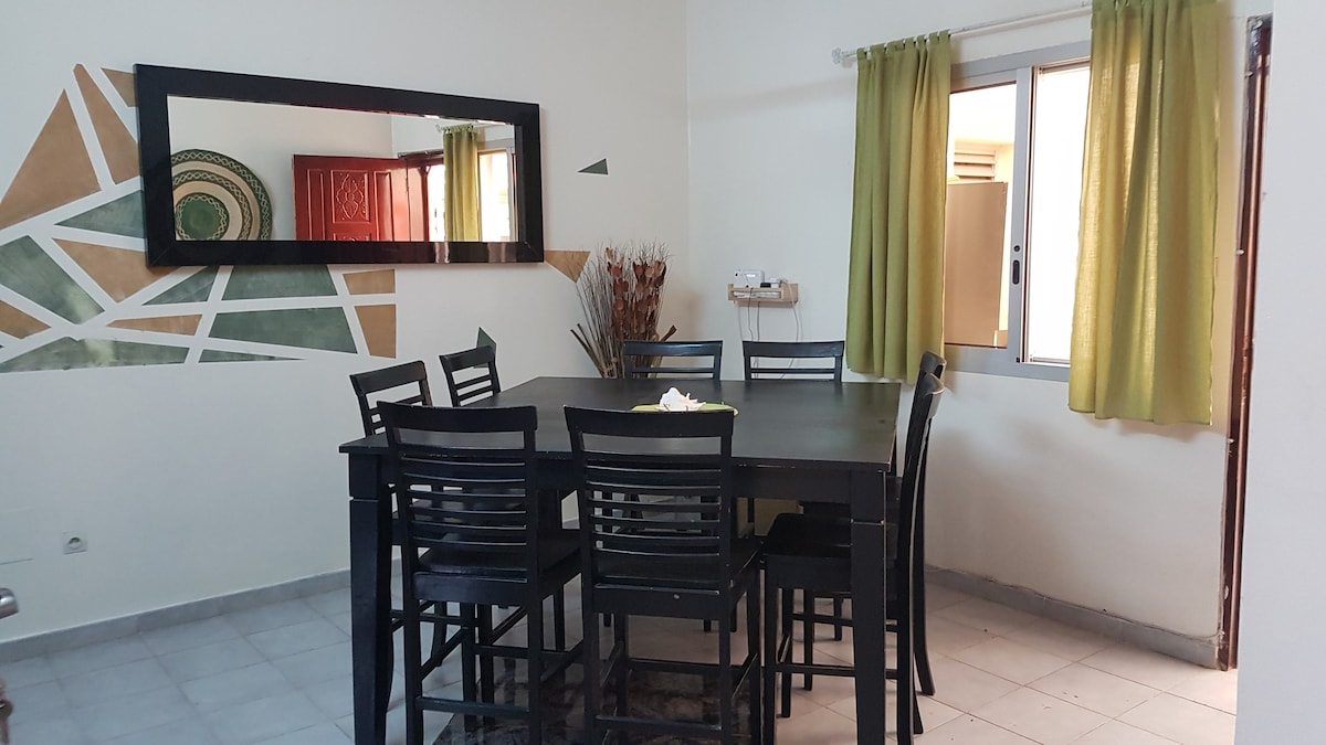 A dining area featuring a large black table surrounded by eight high-backed chairs. A decorative mirror enhances the space along one wall, while green curtains frame the window, allowing natural light to illuminate the room. Subtle geometric patterns are displayed on the adjacent wall.