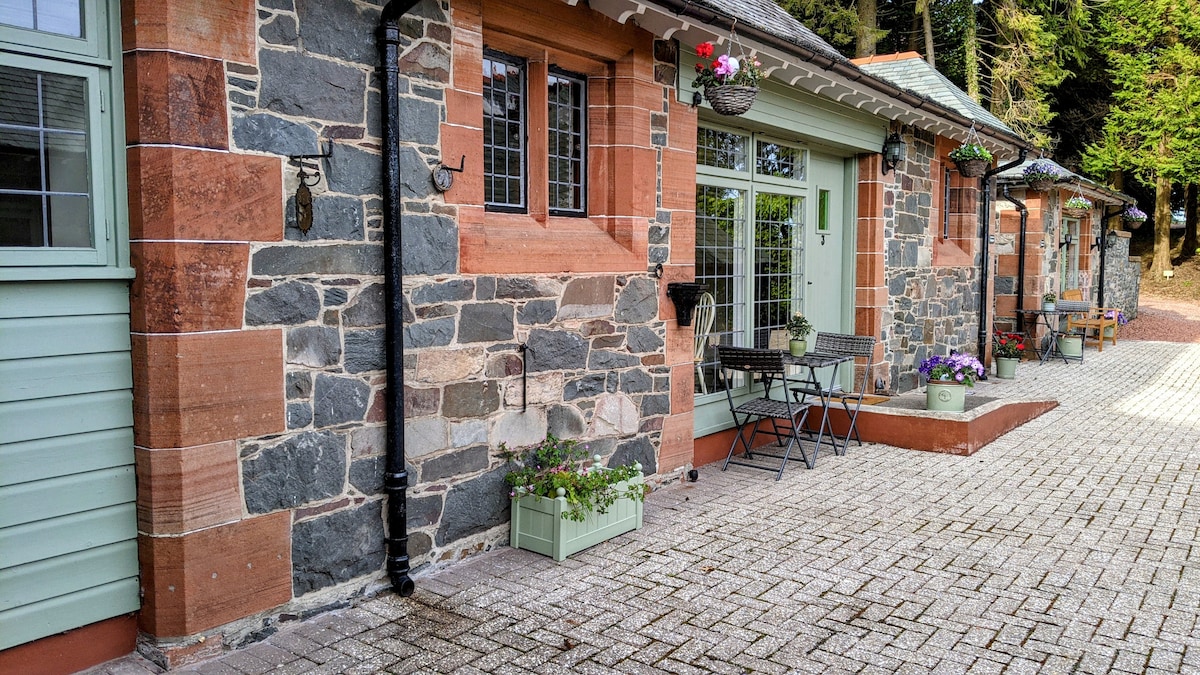 The exterior of the cottage is characterized by a blend of stonework and vibrant wood accents. A paved pathway leads to large windows, allowing natural light inside. Flower boxes adorn the entrance, and outdoor seating is provided with a small table and chairs, enhancing the tranquil setting.