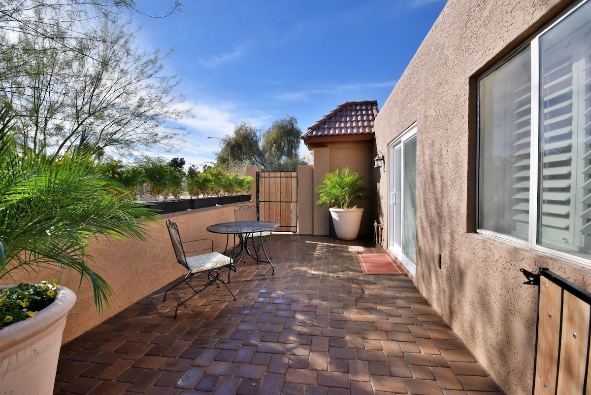 An outdoor patio area is featured, with a tiled floor and a small round table accompanied by two chairs. Lush greenery is present in planters, along with desert landscaping in the background. A gate provides access to the space, with a clear blue sky above.