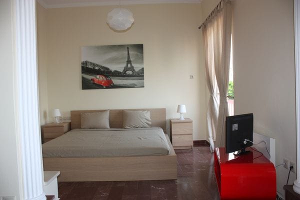 A bedroom features a spacious bed with neutral bedding, complemented by bedside tables and lamps. A television is positioned on a bright red console. A wall artwork depicts the Eiffel Tower, and large windows allow natural light to fill the room, framed by sheer curtains.