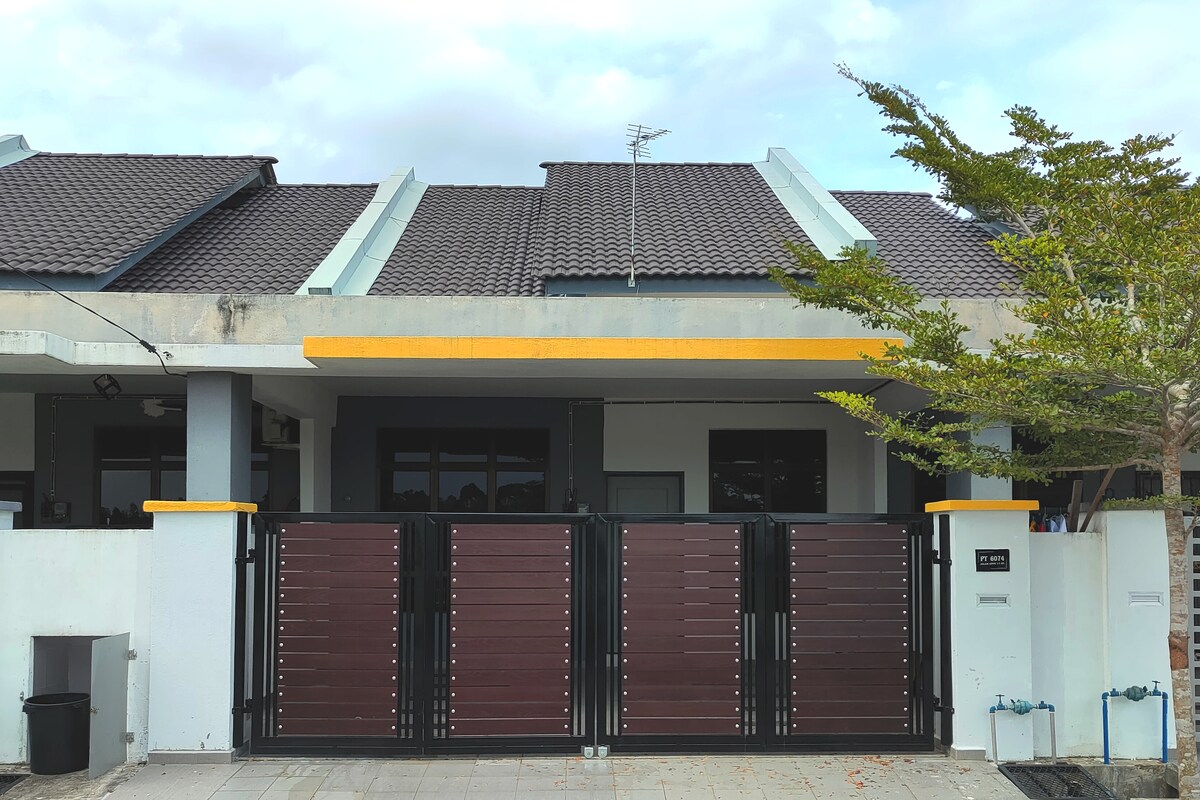 The exterior of the house features a modern design with a combination of muted colors. A secured entrance is marked by a large, dark gate with horizontal slats, complemented by a light-colored facade and a landscaped area with greenery.