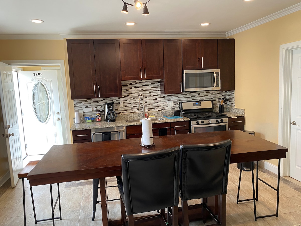 A spacious kitchen area features dark wood cabinetry and granite countertops. A sizable table with black chairs is positioned in the center. Stainless steel appliances include a microwave and a stove, with decorative tile backsplash adding a modern touch. Natural light enters through the entrance door.