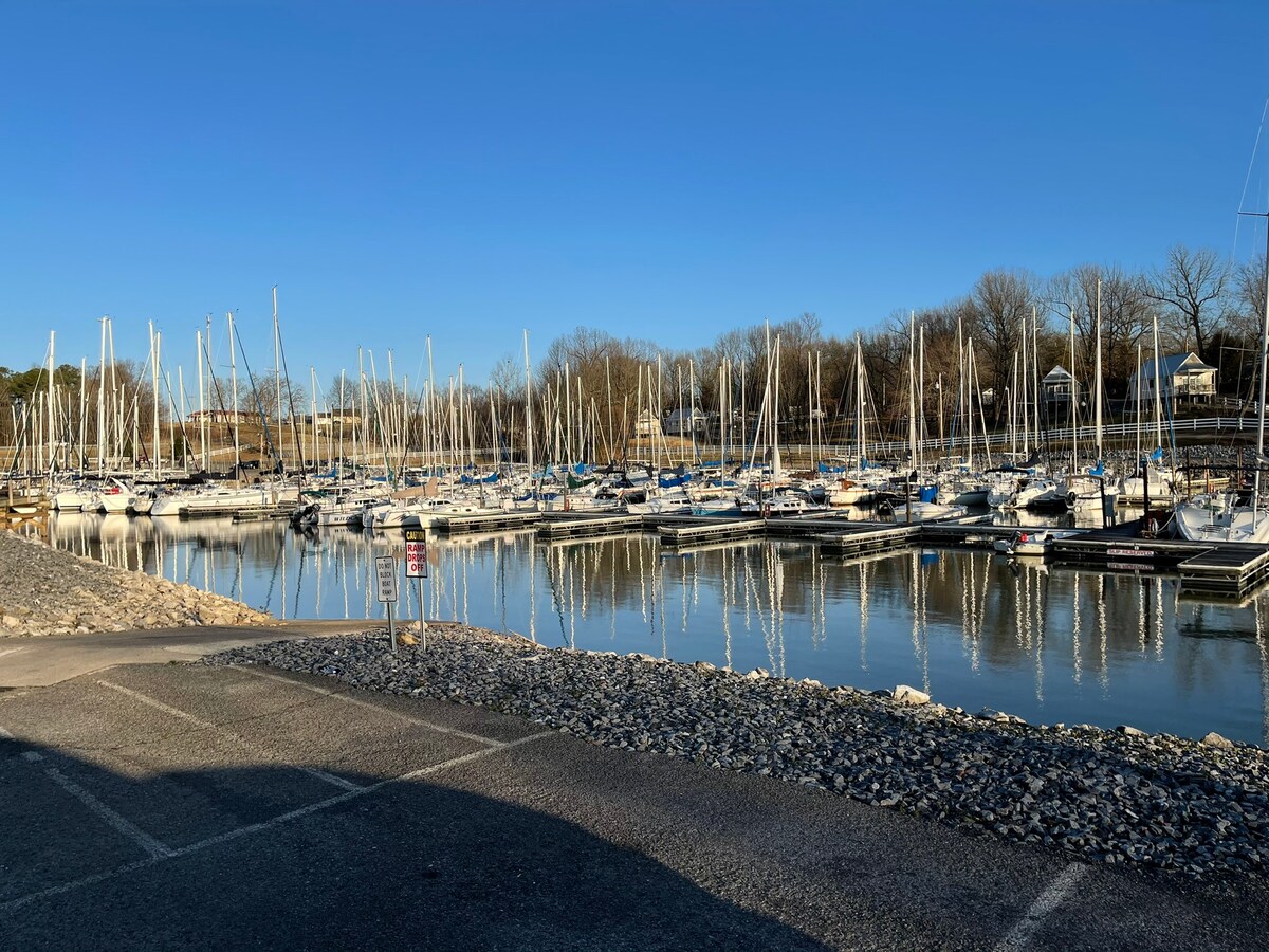 A serene marina is depicted, featuring numerous sailboats docked in calm waters. The scene is framed by clear blue skies and trees lining the shore, with gentle reflections visible on the water's surface. Parking is available nearby, with a gravel pathway leading to the docks.