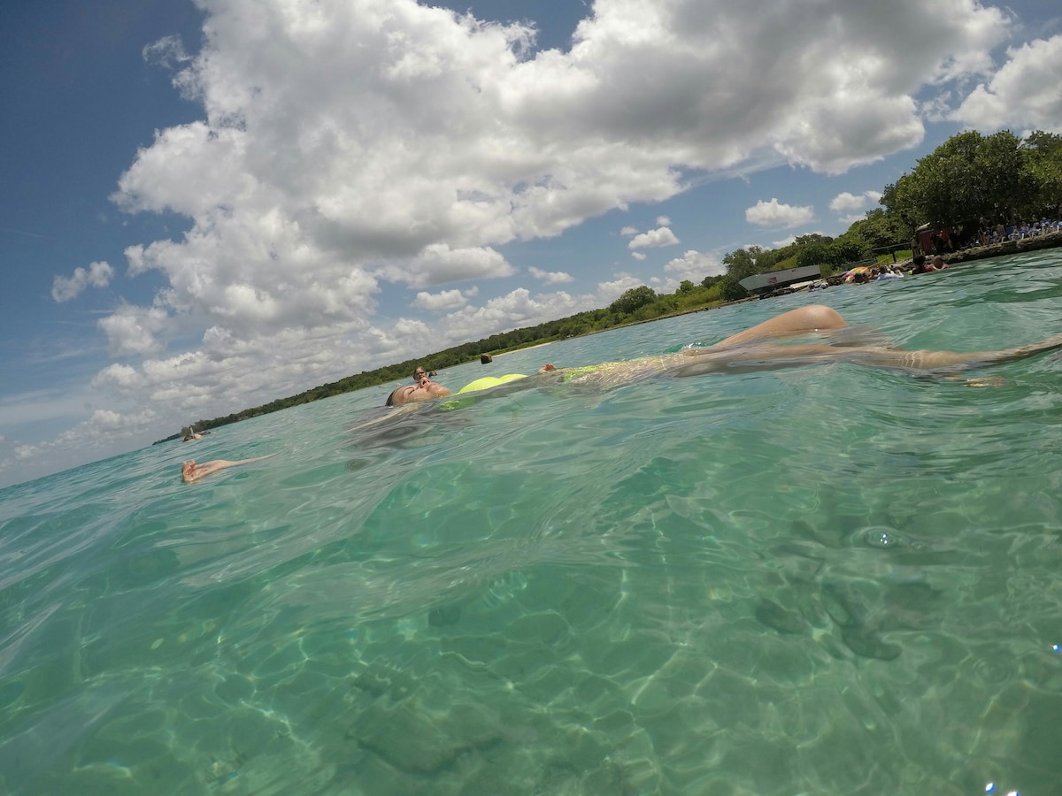 Clear turquoise waters are depicted, with a person floating peacefully on the surface. Bright clouds drift across a blue sky, while lush greenery outlines the shoreline. The scene conveys a serene environment, ideal for snorkeling and enjoying the natural surroundings.