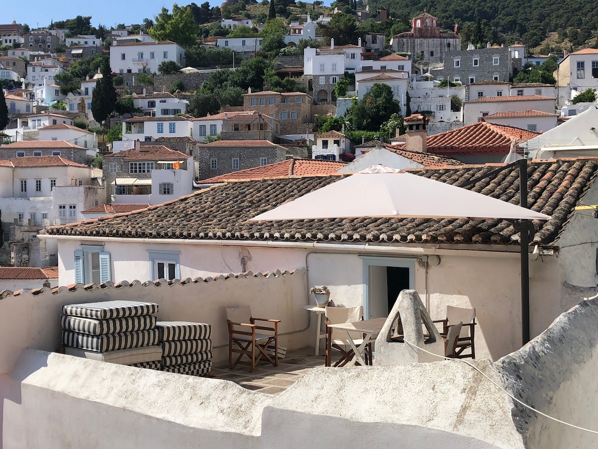 A terrace is seen, featuring a table and chairs under a large umbrella, with a backdrop of traditional houses on the hillside. Textured stone walls and terracotta rooftops create an inviting outdoor space, emphasizing the surrounding picturesque landscape.