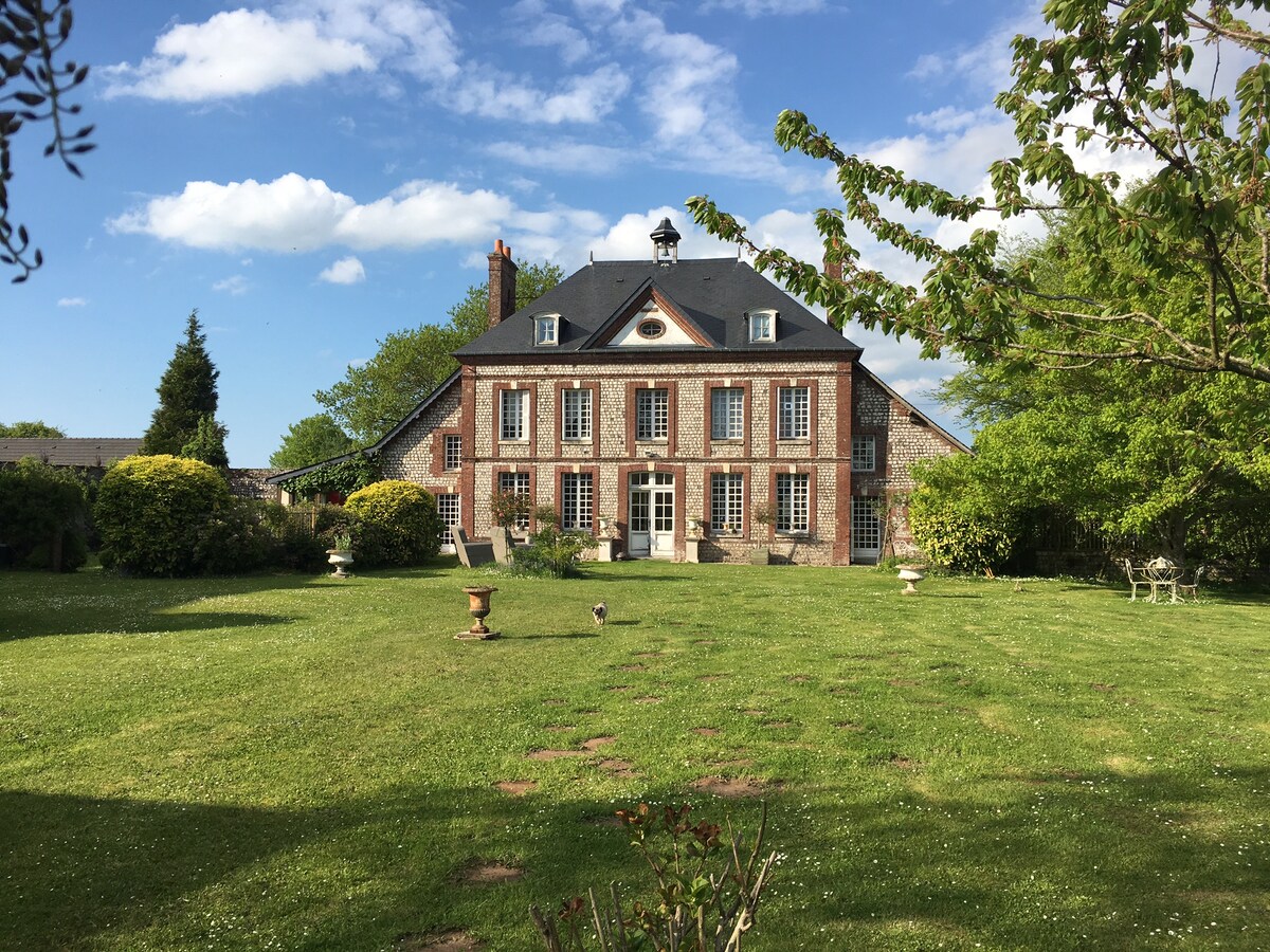 A two-story house with a brick facade and a slate roof is set amidst a generous green lawn, bordered by trees and flowering shrubs. The exterior features multiple windows and a central entrance with double doors, contributing to a spacious outdoor ambiance.