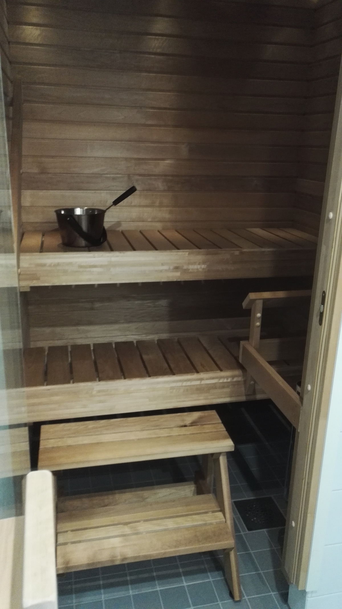 A wooden sauna interior is shown, featuring tiered benches made of light-colored wood. A small wooden stool is positioned near the entrance, while a metal bucket and ladle sit on the upper shelf, indicating a cozy spot for relaxation.