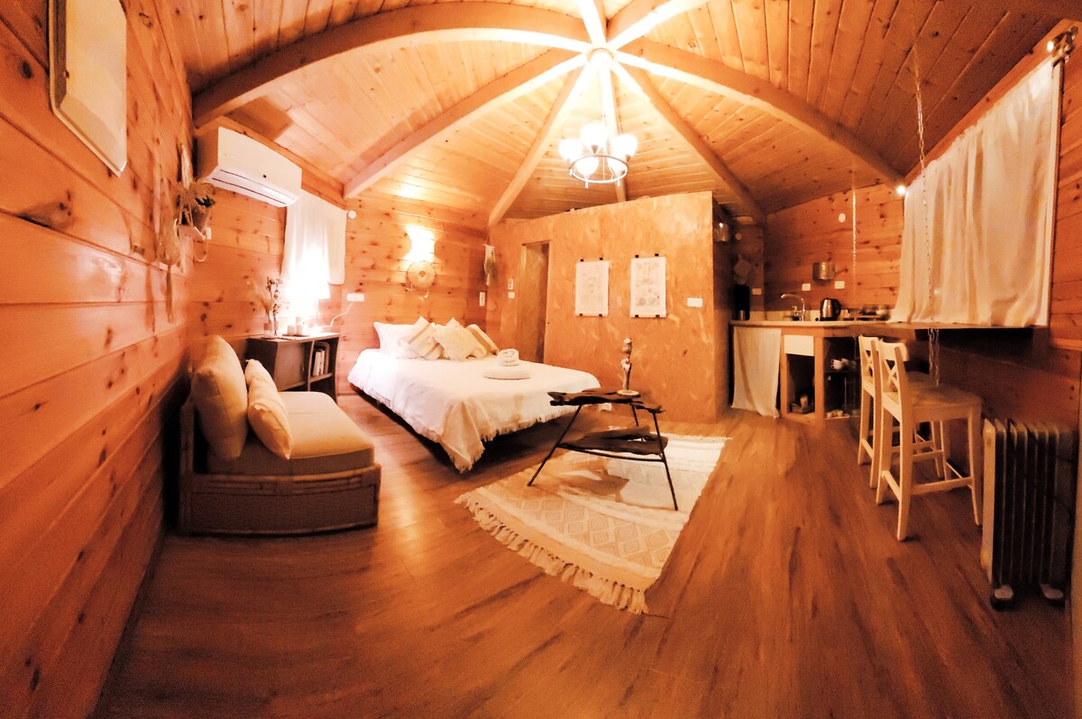 A wooden cabin interior is displayed, featuring a spacious bed with soft linens and a cozy seating area. The ceiling boasts wooden beams, and warm lighting enhances the inviting atmosphere. A small kitchenette is visible, along with a dining table and chairs, contributing to the overall comfort.