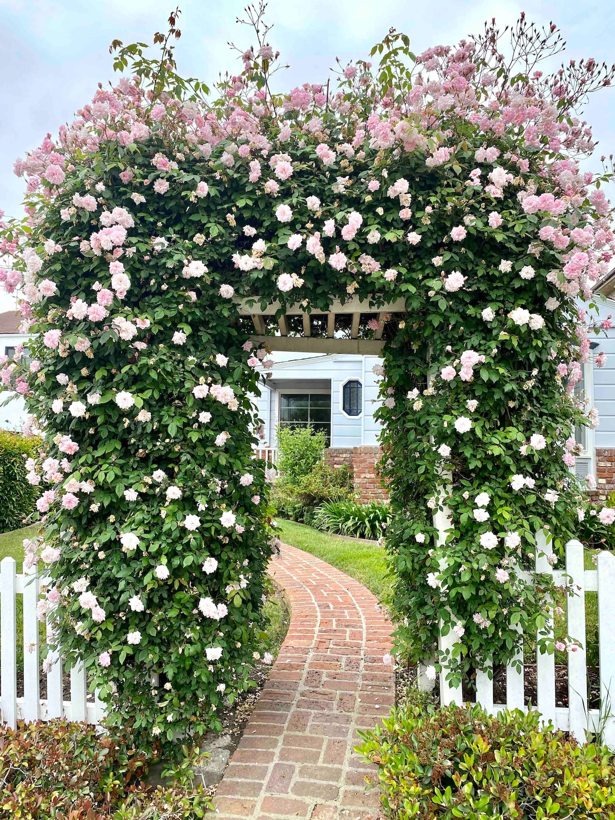 An arched trellis covered in blooming pink roses frames a winding brick pathway. The garden is bordered by a white picket fence, while lush greenery enhances the entrance to the home beyond. The soft colors of the flowers complement the surrounding natural beauty.