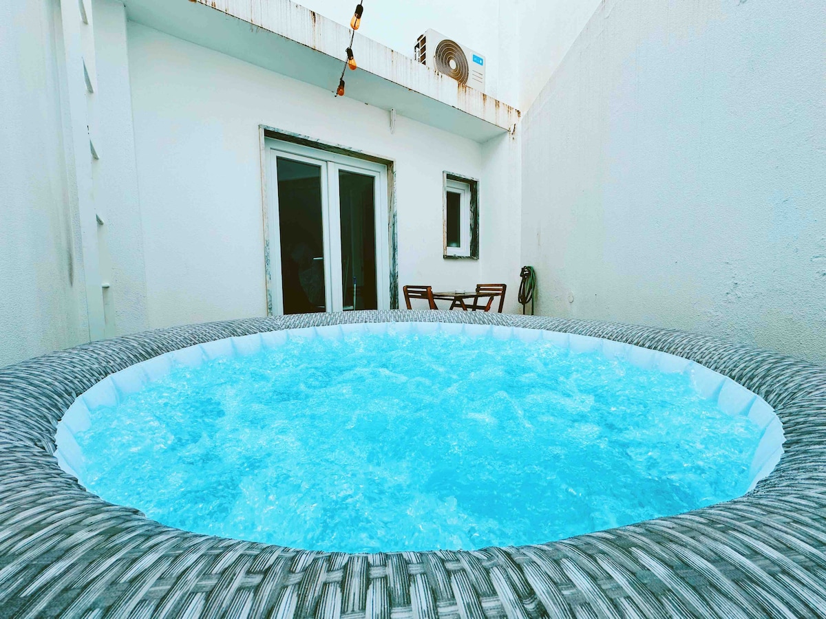 A private jacuzzi with sparkling blue water is set against a light-colored wall, creating a serene atmosphere. Two wooden chairs are positioned nearby, inviting relaxation. Soft lighting strings are visible above, adding a gentle ambiance to the outdoor space.