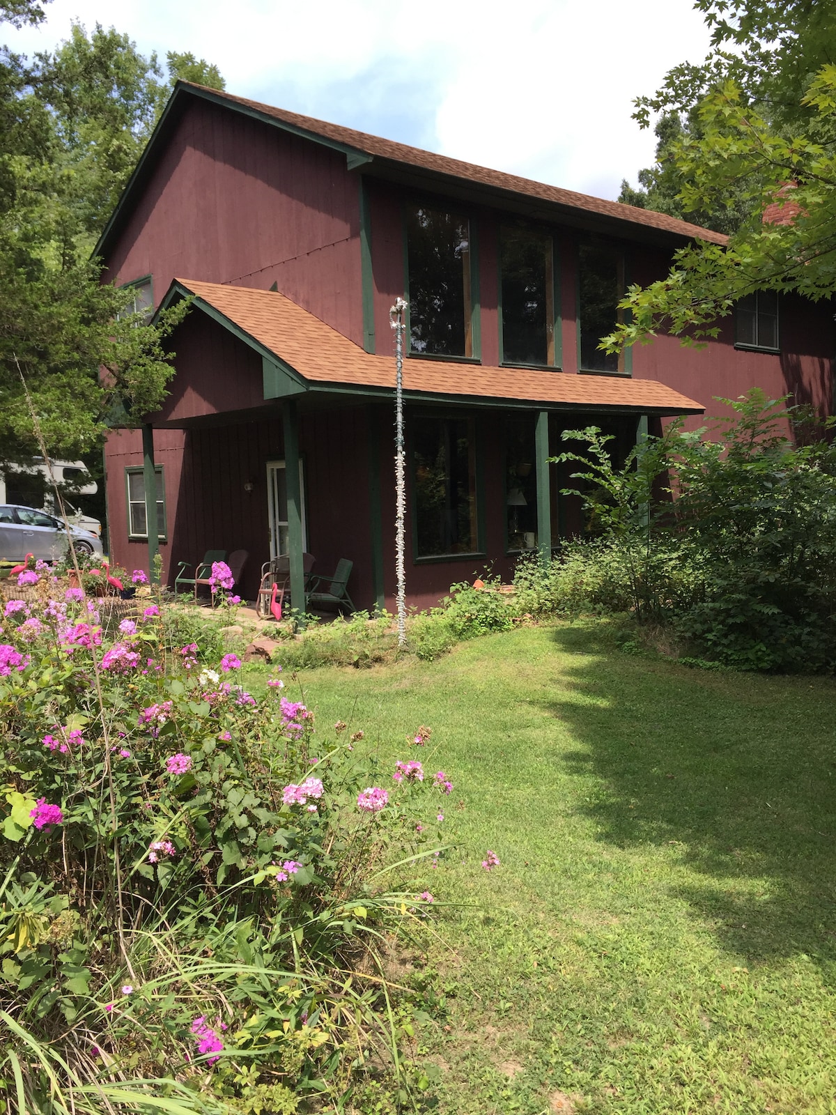 A rustic two-story house, painted in rich maroon, is surrounded by lush greenery and colorful flowers. Large windows allow natural light to illuminate the interior. An inviting front porch features seating, providing a tranquil view of the garden and the peaceful country setting.