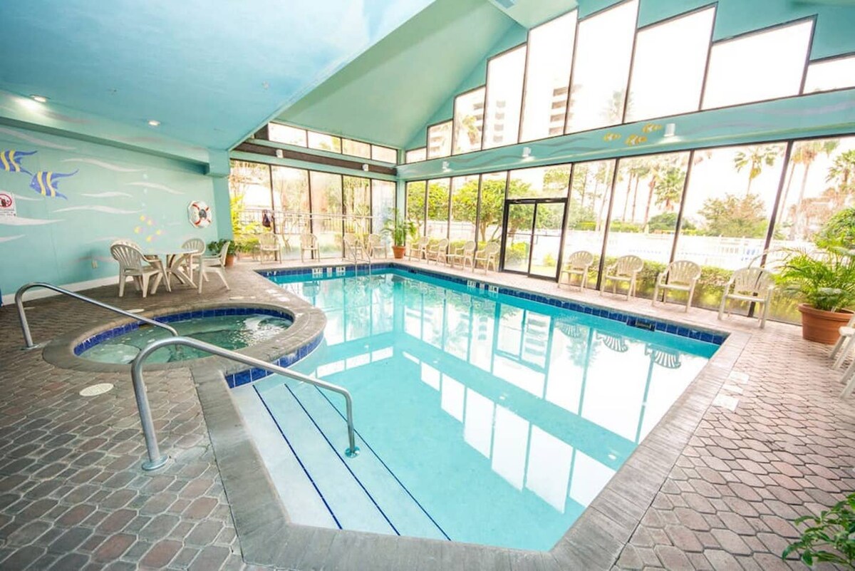 An indoor heated pool area features large windows providing ample natural light and views of the outdoor landscape. A connected jacuzzi sits alongside the pool. Several lounge chairs are arranged around the pool's perimeter, and vibrant wall art enhances the space.