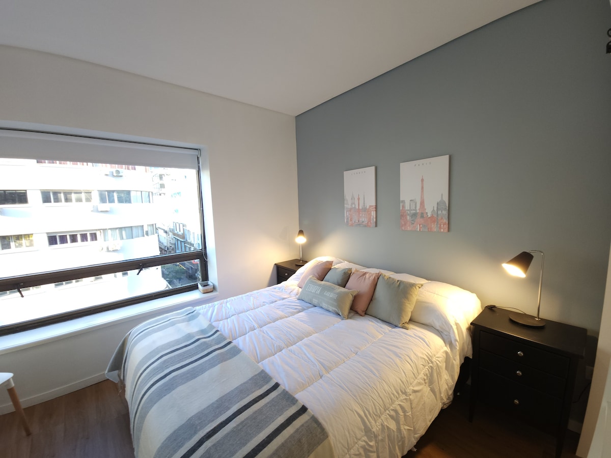 A bright sleeping area features a spacious bed with soft bedding, complemented by decorative pillows. Two wall art pieces adorn the light-colored wall above the bed. Natural light enters through a large window, and bedside tables with lamps are positioned on either side.