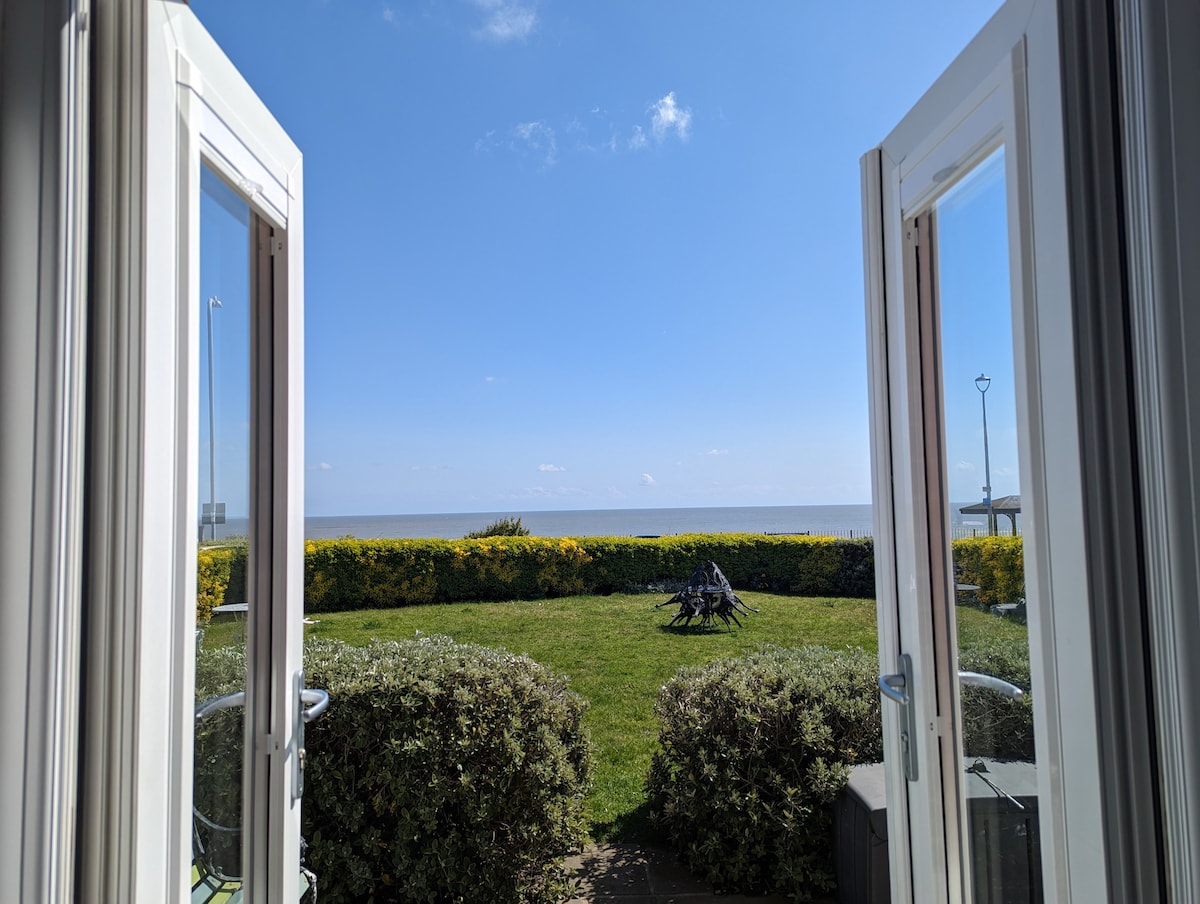 French doors are open, revealing a clear blue sky and a view of the sea. Lush greenery frames the outdoor space, which includes a well-maintained lawn and seating area, while the gentle horizon can be seen in the distance.