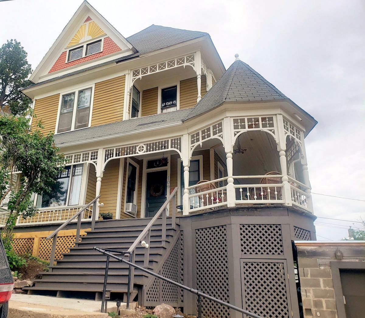 The exterior of a restored Victorian home features a vibrant yellow facade and ornate architectural details. A spacious porch wraps around the front, supported by decorative spindles. The entrance stairs lead up to the main door, framed by lush greenery and a hilly landscape.