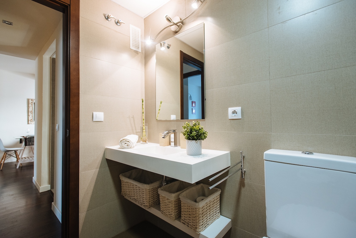 The bathroom features a modern sink with a wide countertop and a rectangular mirror above it. Beneath the sink, two baskets provide additional storage. A contemporary toilet is positioned nearby, with neutral-colored tiles adorning the walls, creating a clean and serene environment.