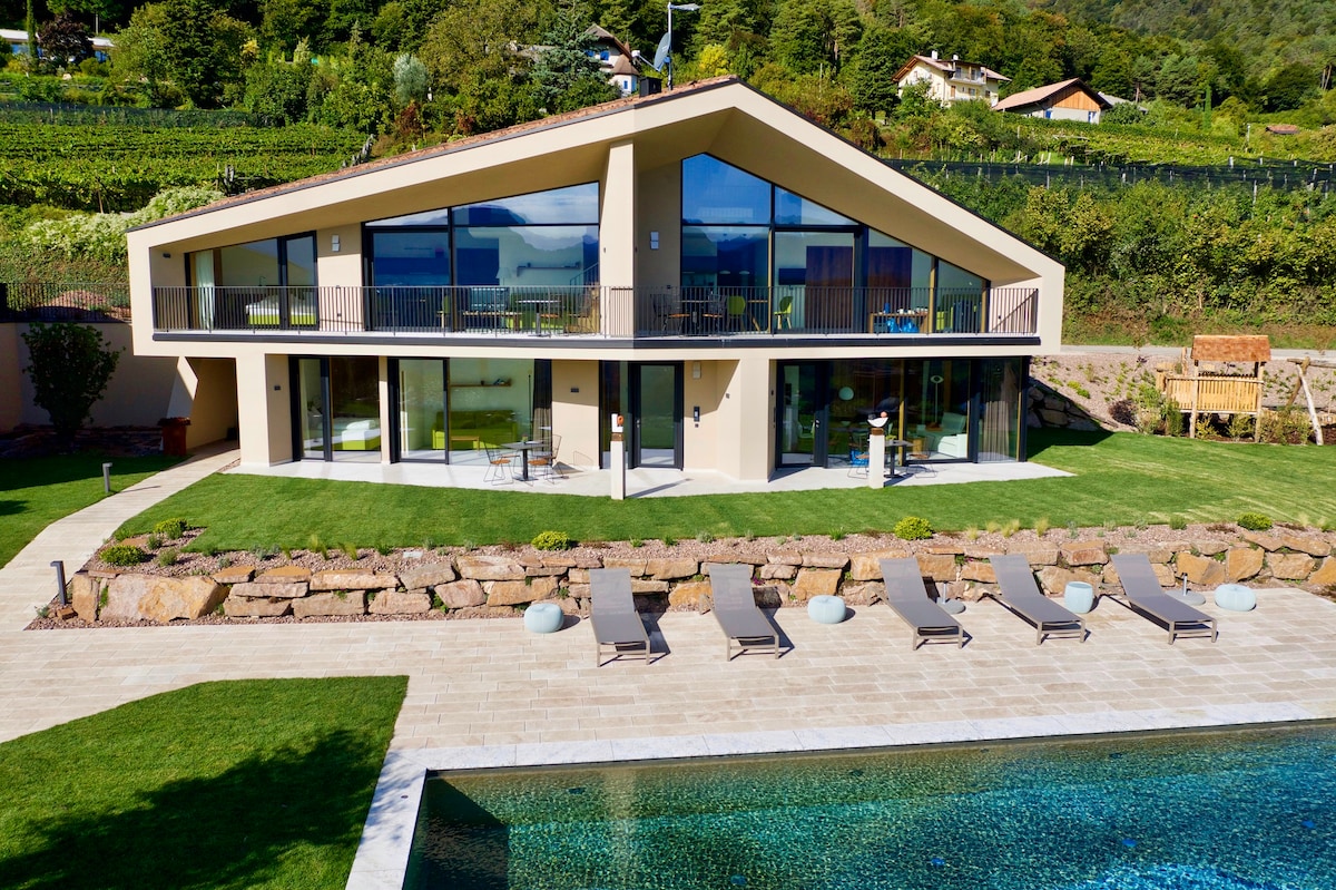 A modern architectural design is featured, showcasing a spacious exterior with large glass panels that provide views of the surrounding vineyards. A well-maintained lawn leads to a swimming pool with lounge chairs positioned nearby, inviting relaxation and enjoyment of the landscape.