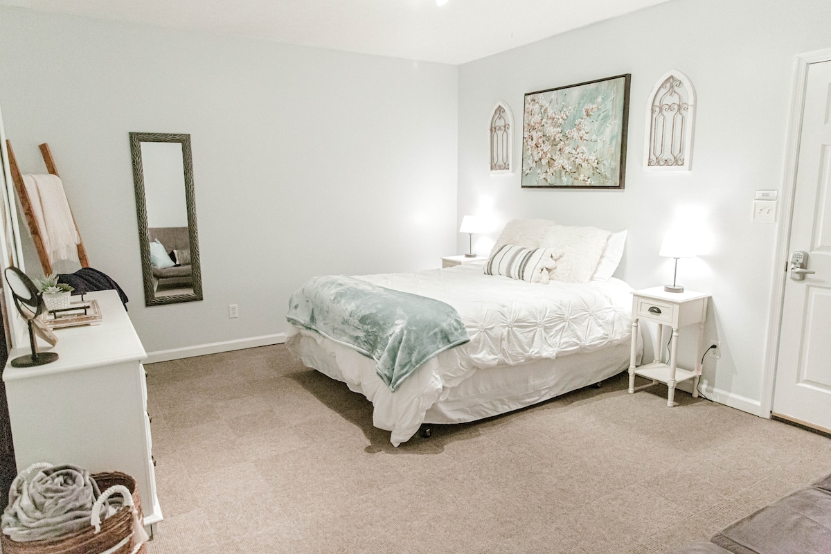 A spacious bedroom is presented with a large bed featuring a soft, light-colored duvet. Nightstands with simple lamps are positioned on either side. A mirror and decorative artwork add subtle elegance to the calming color palette of light grey walls.