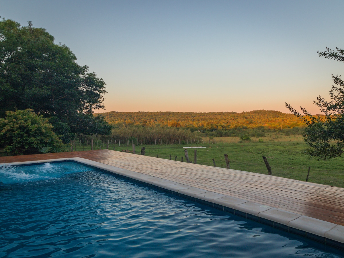 A swimming pool is featured with serene water reflecting the evening light, bordered by a wooden deck. Beyond the pool, lush greenery and rolling hills are visible, adding to the tranquil atmosphere of the surrounding nature.